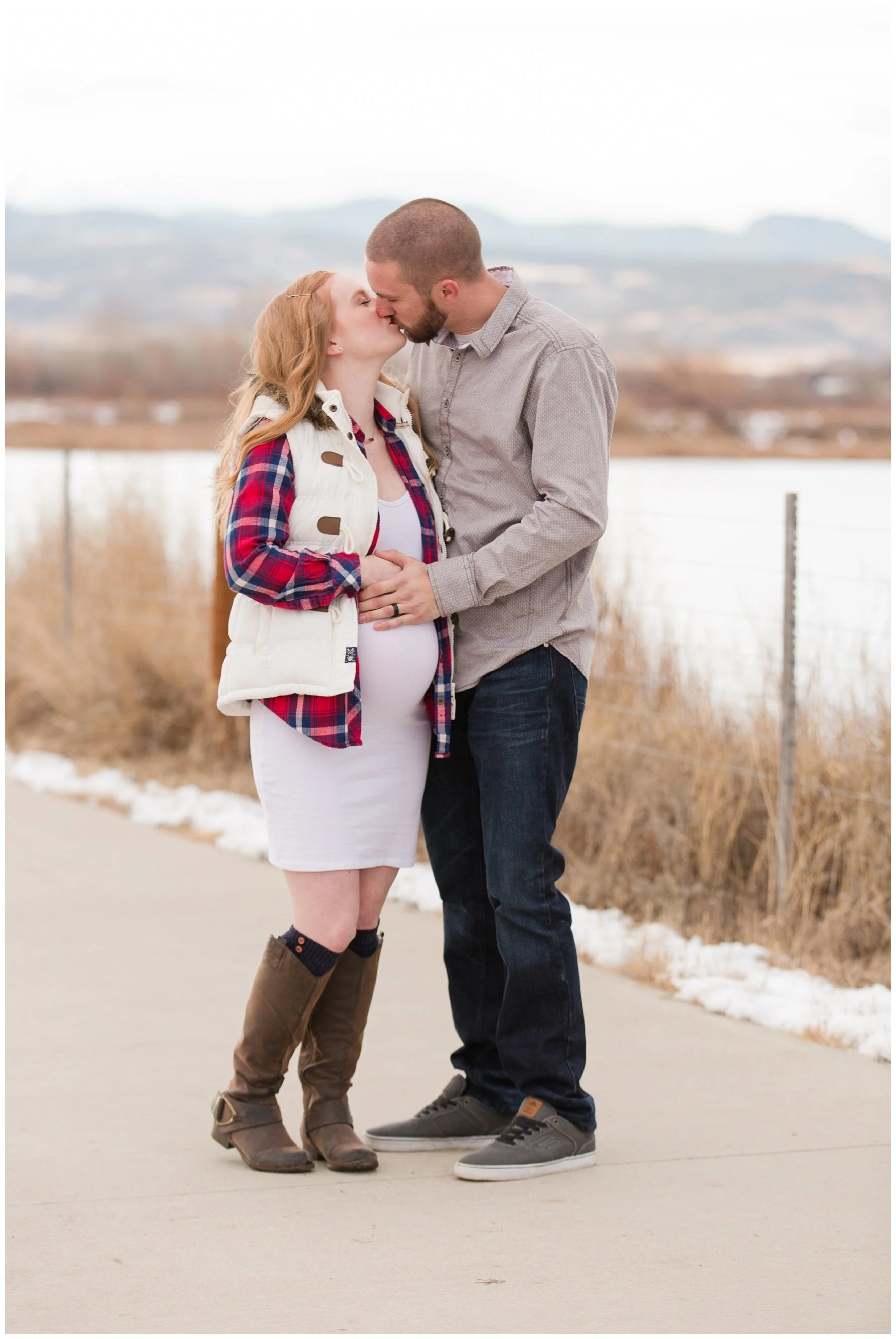A pregnant woman and a man standing close together on a sidewalk, sharing a kiss outdoors, with a rural landscape and overcast sky in the background.