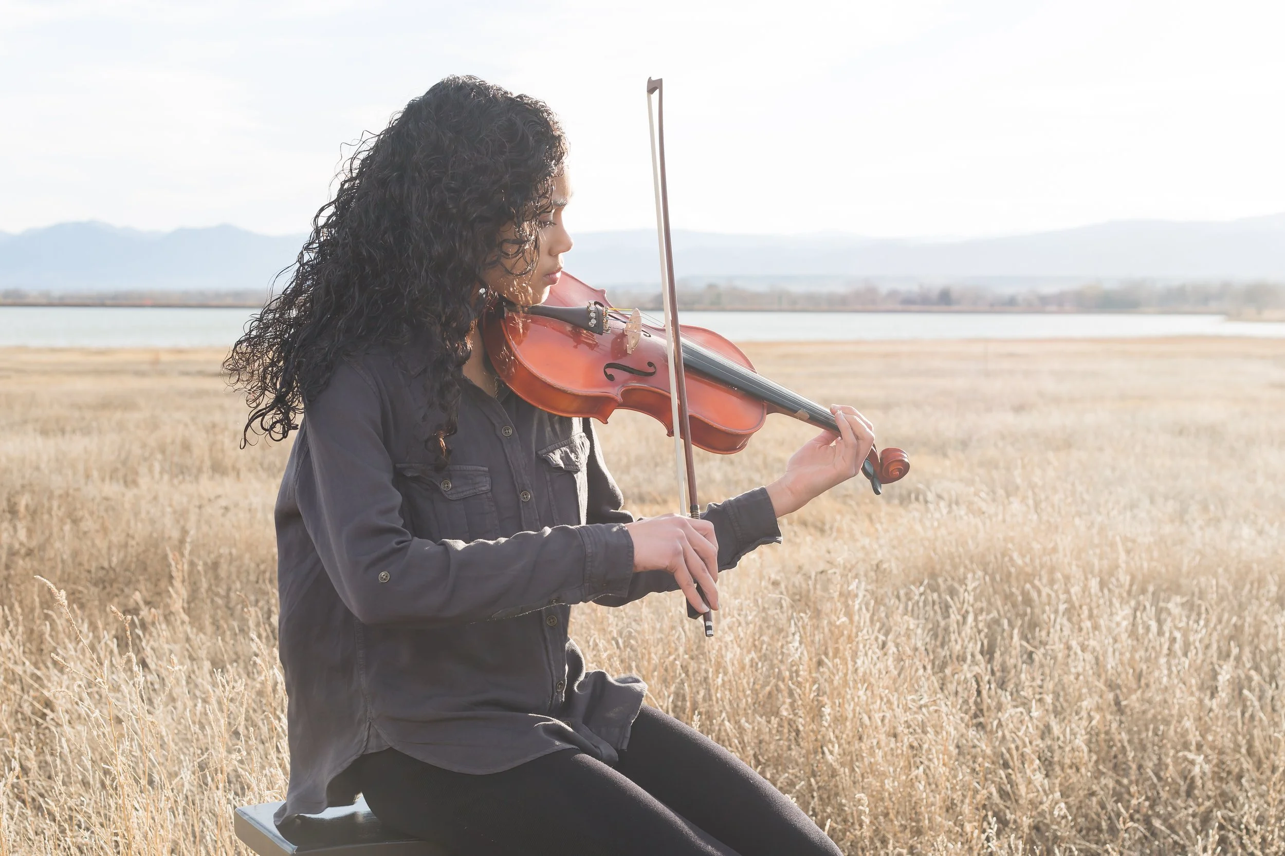 A dramatic fine art senior portrait session featuring a girl playing the violin outdoors in a field of tall golden grass with mountain views at McIntosh Lake in Longmont, Colorado.