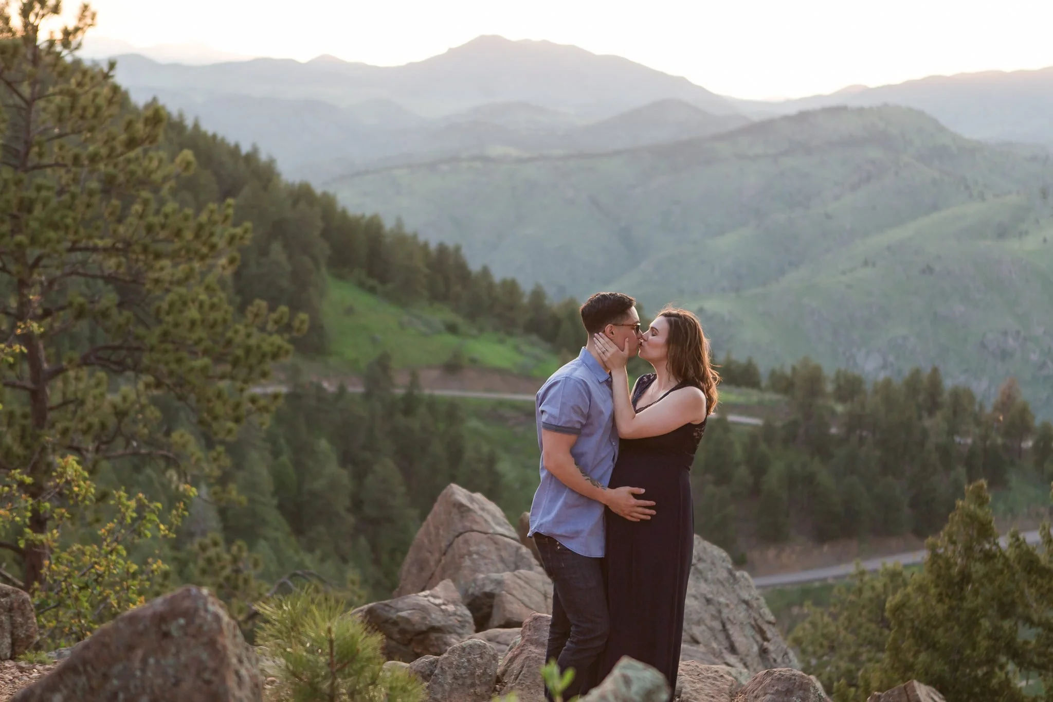 A couple kissing outdoors on a rocky area with mountains and trees in the background during sunset.