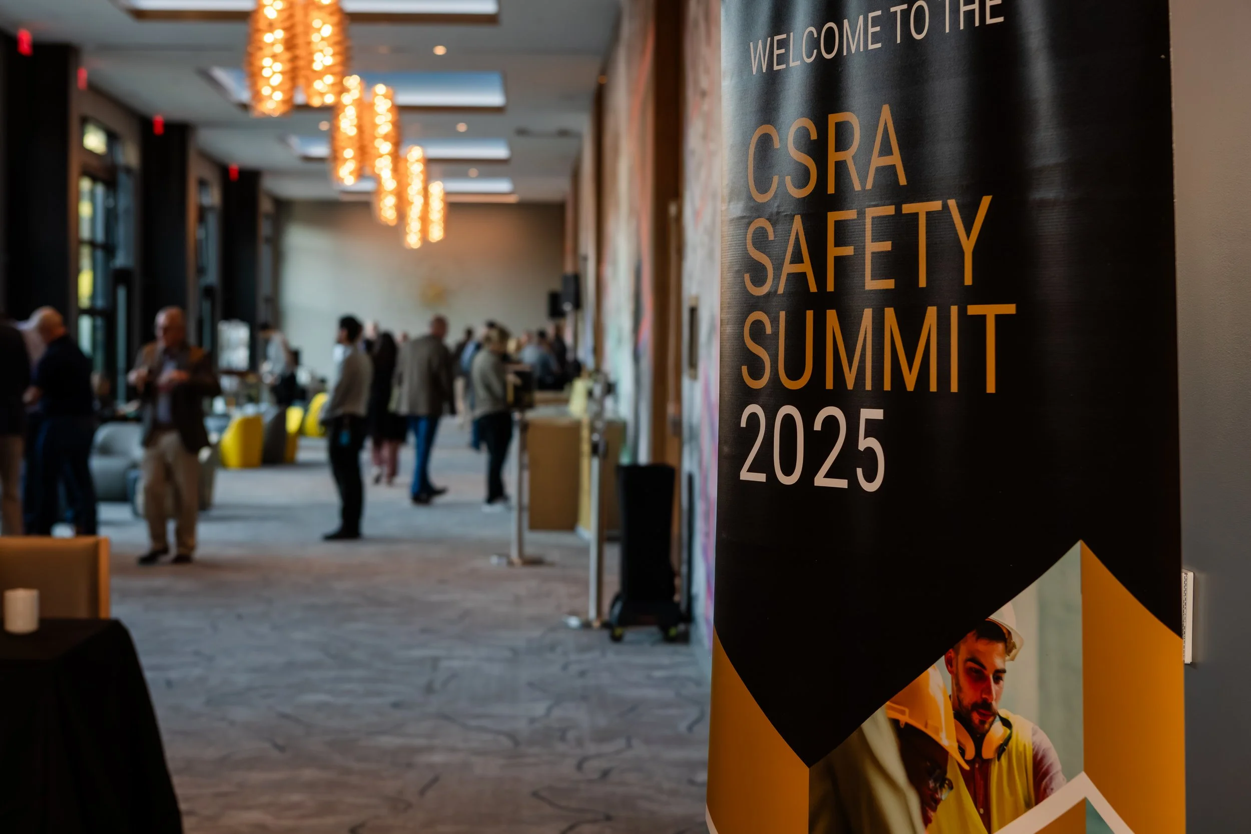 A black vertical banner with orange text reading "Welcome to the CSRA Safety Summit 2025" is displayed at the event. The background shows an indoor conference space with people mingling, some standing near tables and others talking, with decorative h