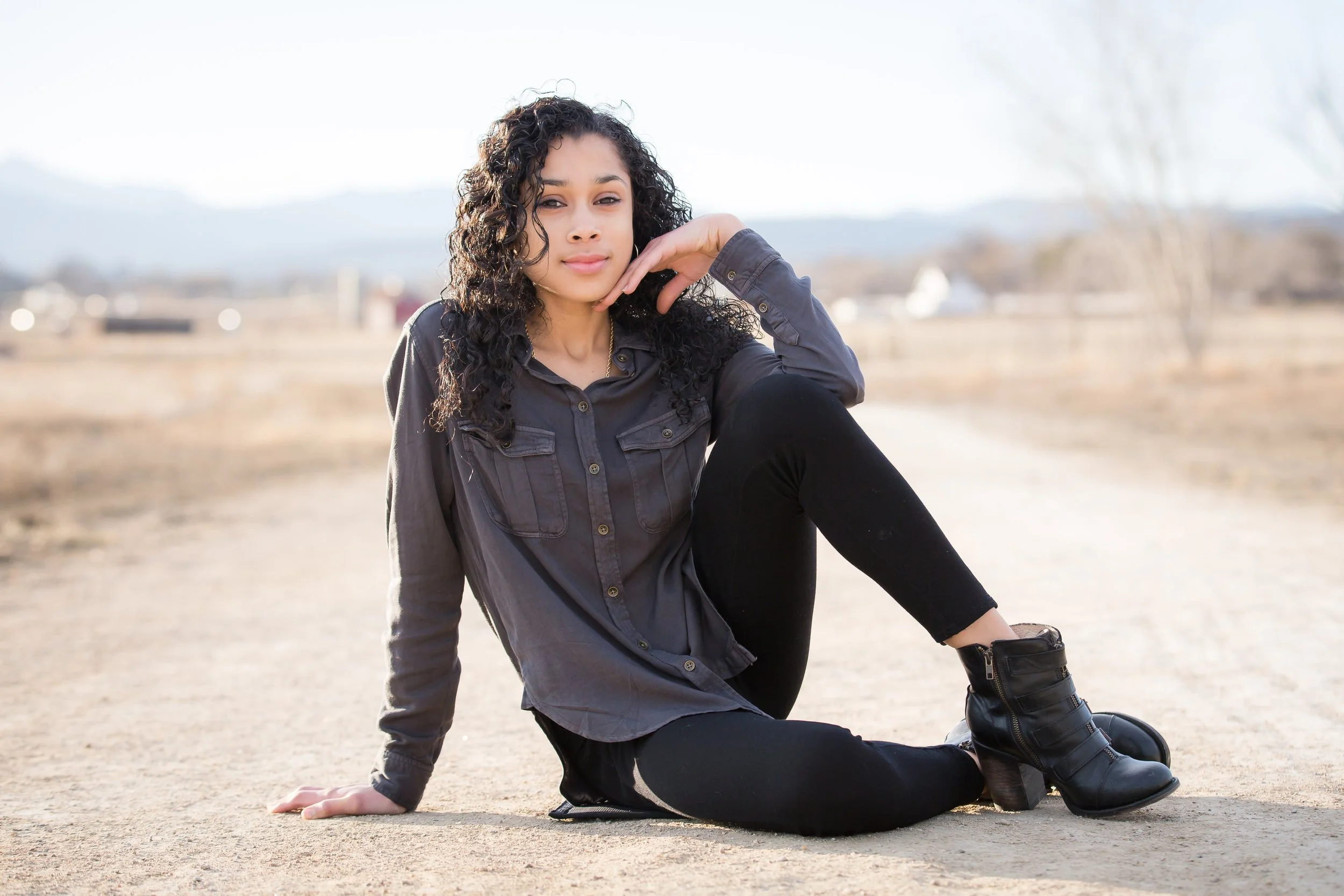 Young woman sitting outdoors on a dirt ground with her left hand resting on the ground, wearing a dark gray military-style jacket, black pants, and black boots, with a blurred natural background and overcast sky.