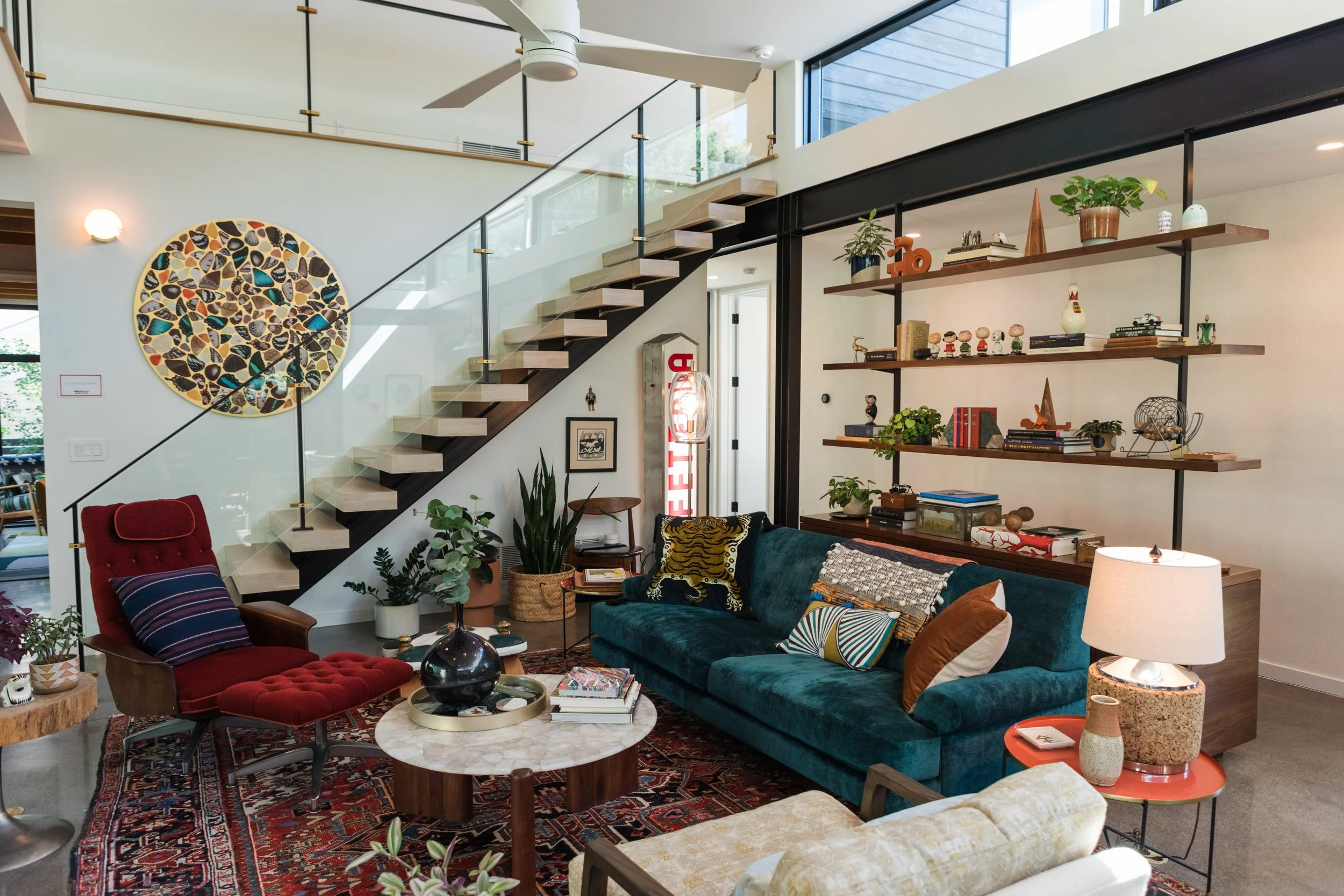 A modern living room with a dark green velvet sofa, a red armchair with a pillow, and a marble-topped coffee table with books and a black vase. There are plants, a patterned rug, and a wooden side table with a lamp. A wall-mounted wooden shelf displa