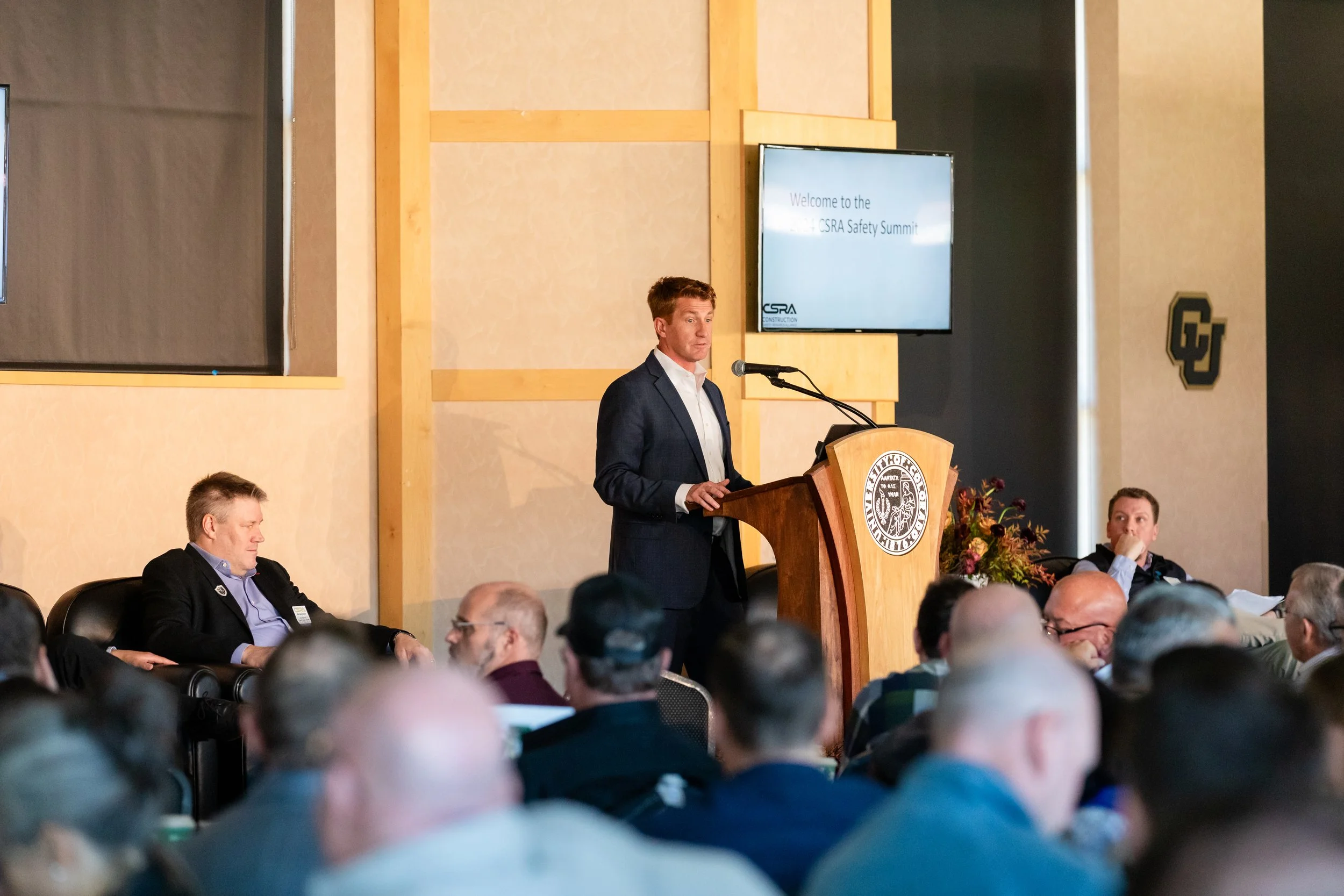 Man in a dark suit speaking at a podium during a safety summit, with attendees seated and listening, and a large screen showing a welcome message behind him.