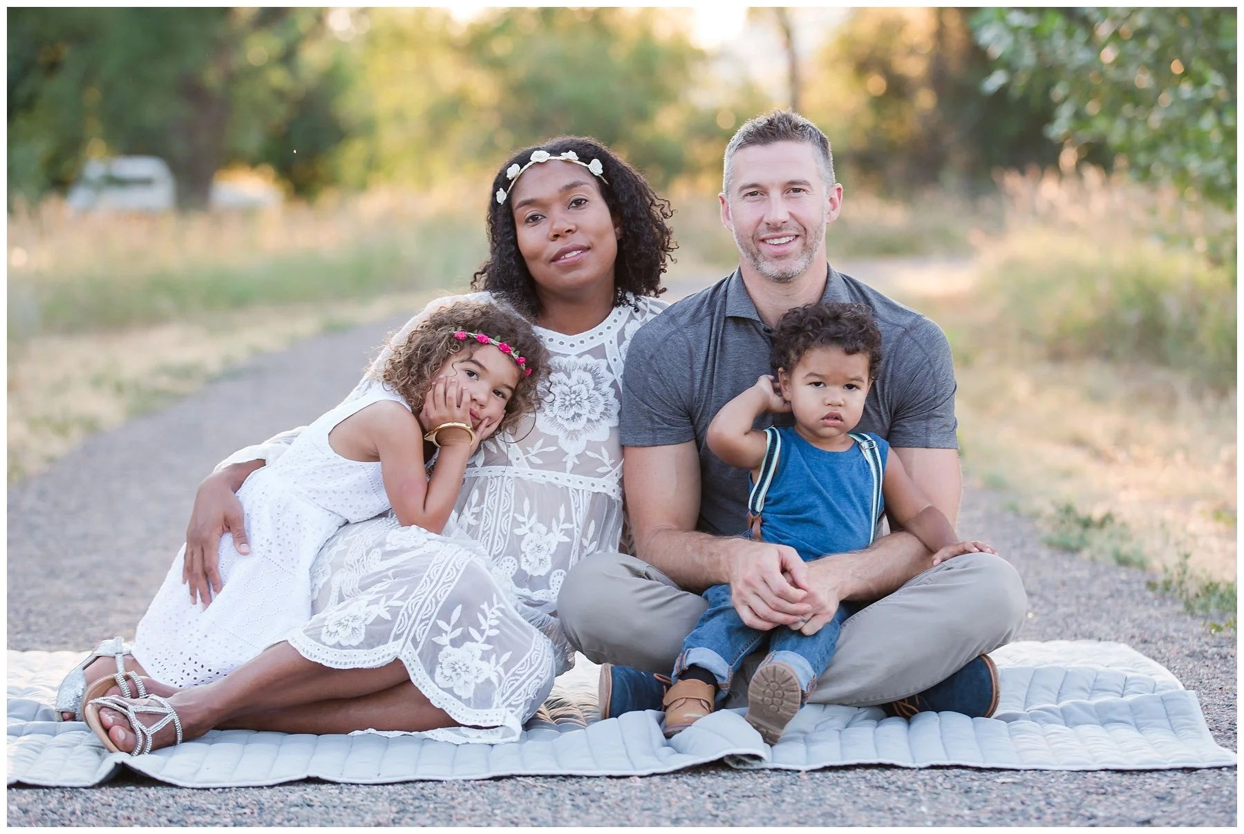 A family of four sitting on a quilt outdoors in a park during fall. The mother has dark curly hair, wearing a white dress with floral embroidery and a white headband. The father has short gray hair and a beard, wearing a gray T-shirt. The young girl 