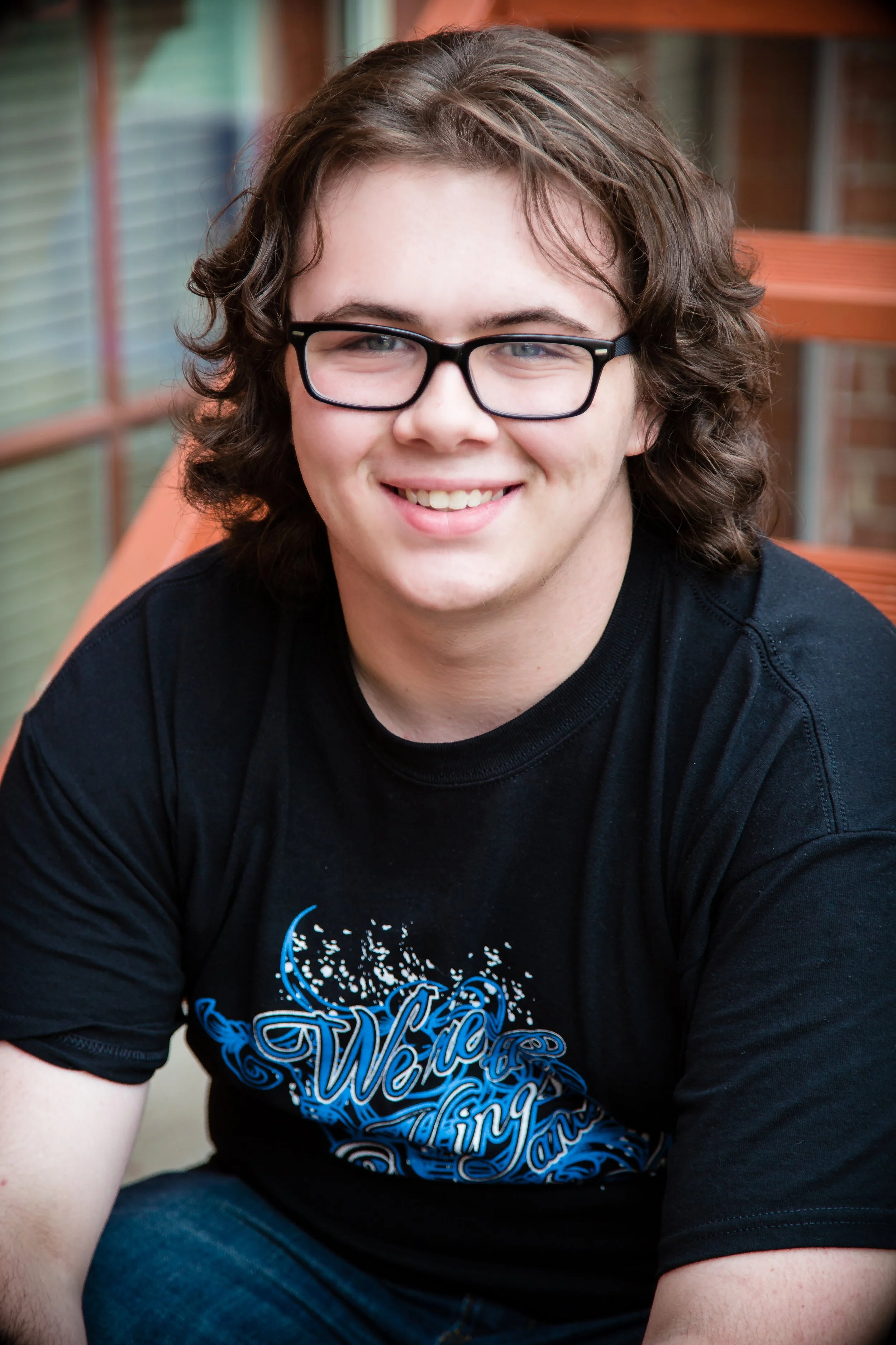 Young man with curly brown hair, glasses, and a black T-shirt with blue and white graphics, smiling and sitting outdoors with a wooden structure in the background.