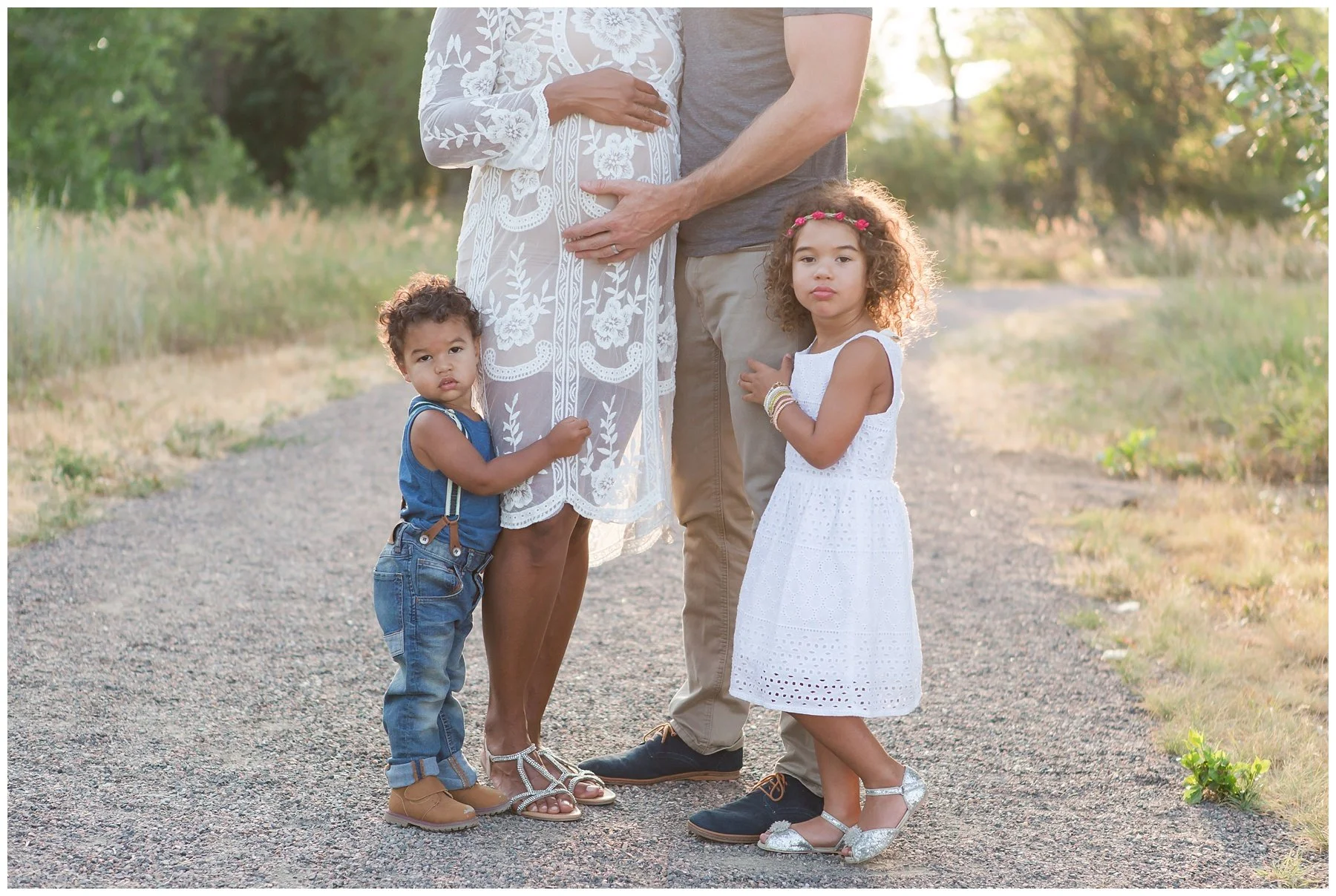 A family of four standing on a gravel path outdoors during sunset. The mother is pregnant, wearing a white lace dress; the father is standing beside her, with one hand on her belly. Two young children, a boy in jeans and a blue shirt and a girl in a 