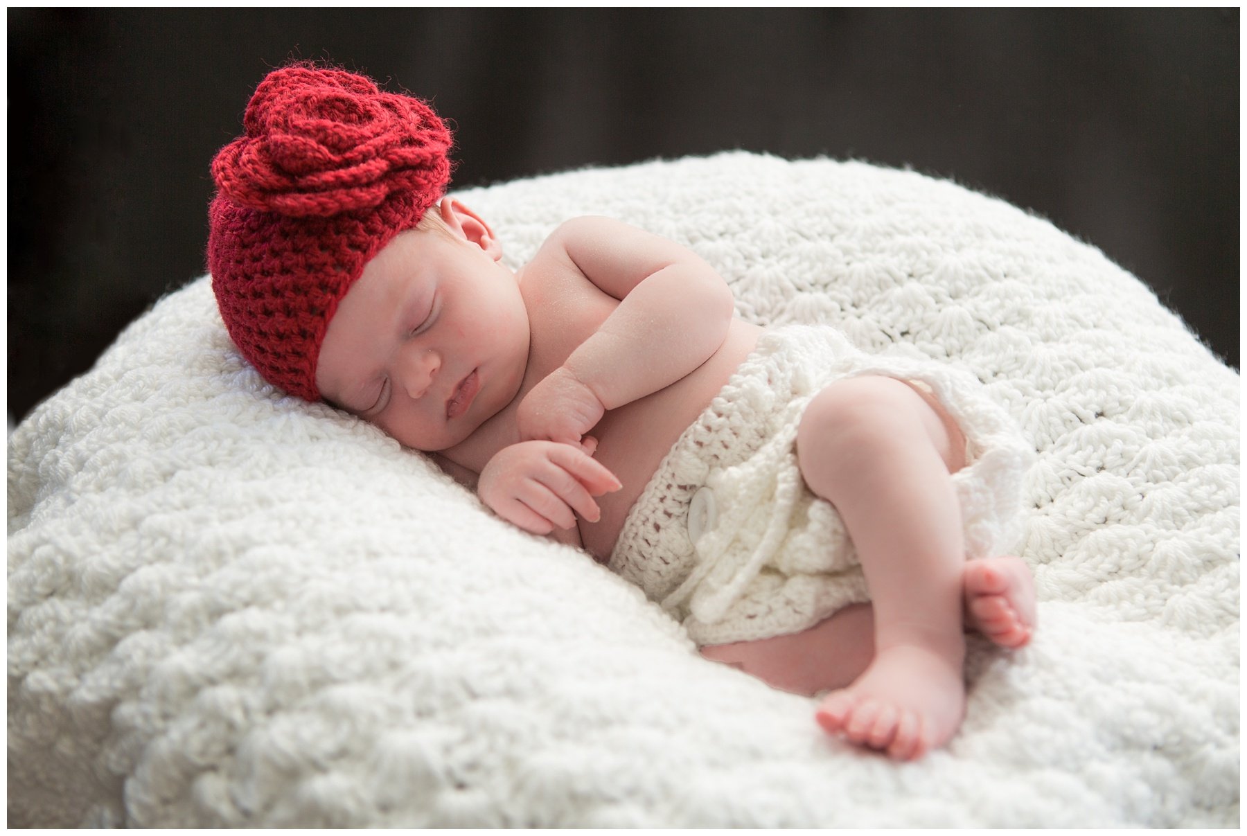 A sleeping baby wearing a red knit hat and cream-colored shorts, lying on a soft, textured white blanket.