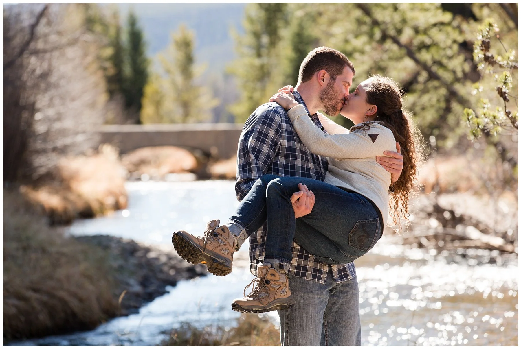 A man carrying a woman in his arms outdoors near a river, with trees and a bridge in the background, on a sunny day.