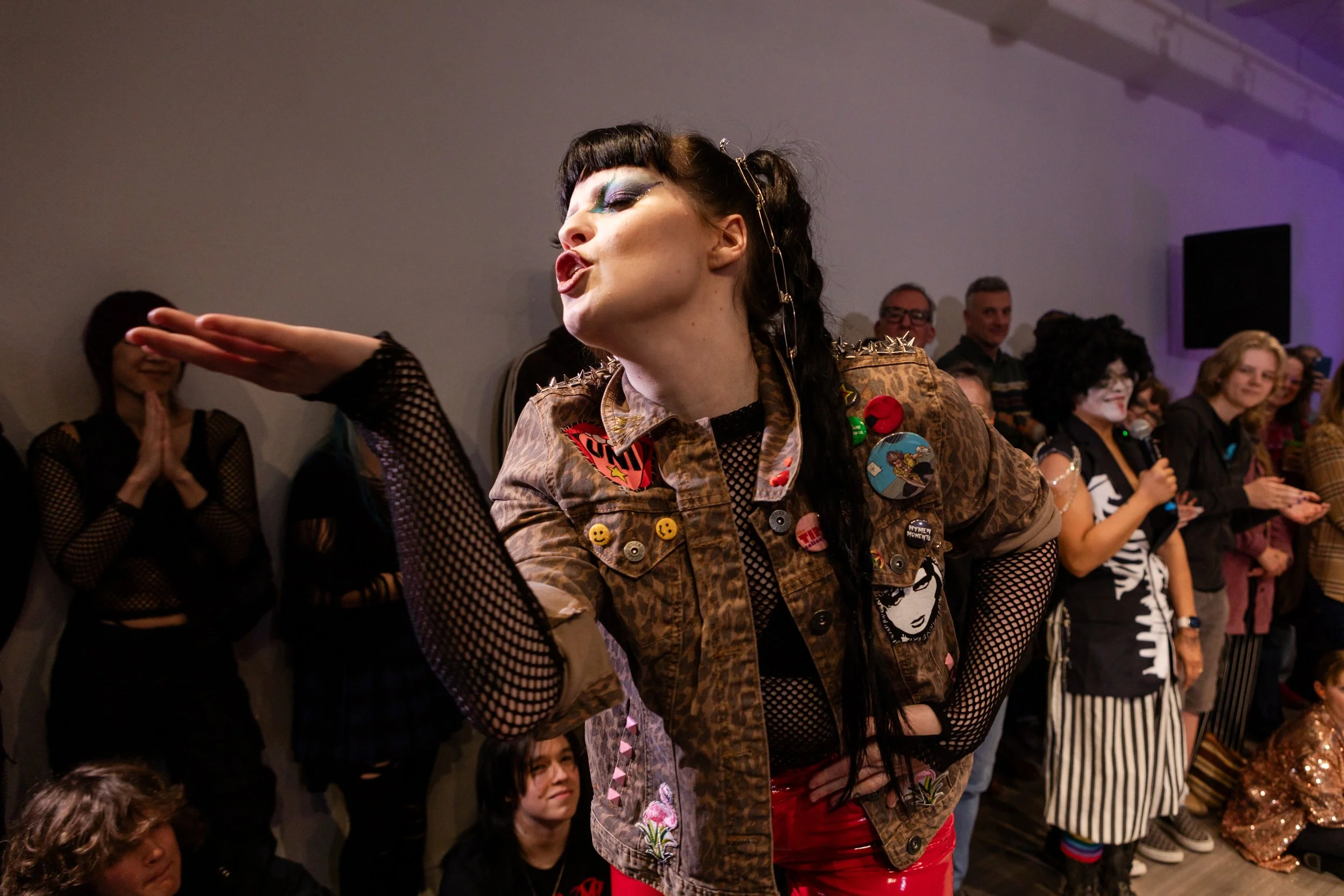 A person with dramatic eye makeup and styled hair blowing a kiss in front of a crowd at a party or gathering.