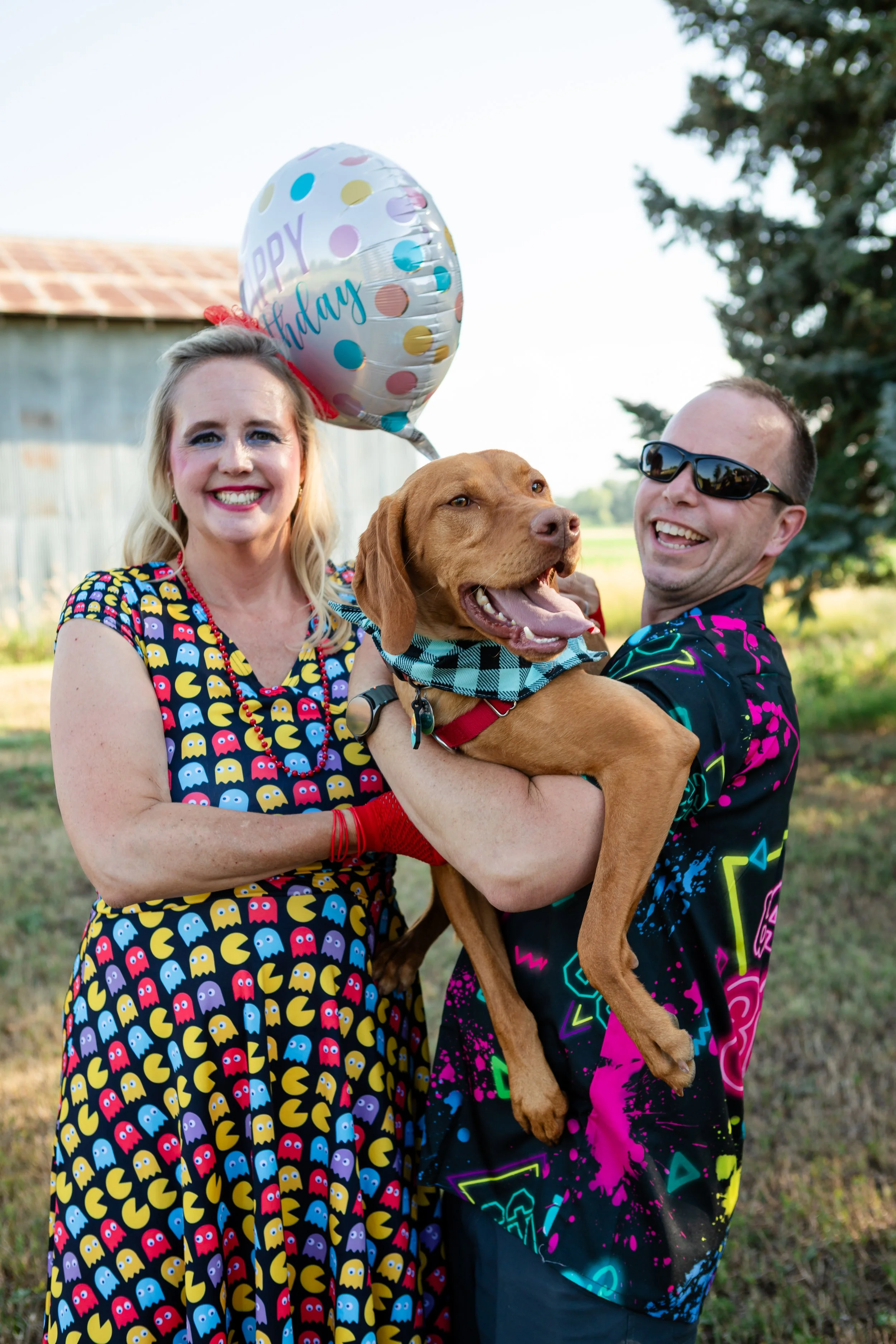 A joyful outdoor portrait of a woman in a retro Pac-Man patterned dress holding her dog during her 50th birthday celebration at Old Elm Farm in Boulder, Colorado.