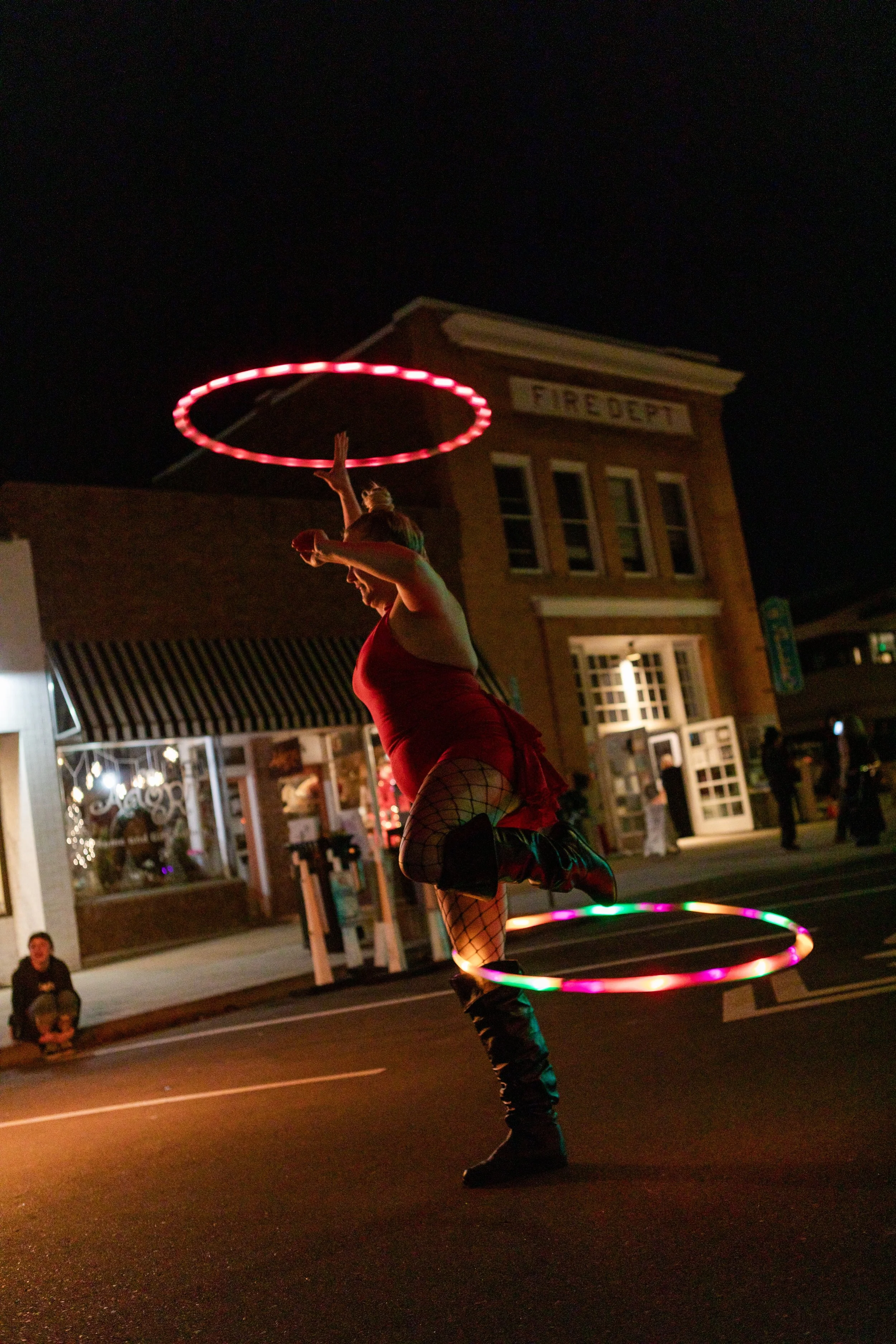 A performer spinning illuminated hula hoops at night in a small town, with buildings and onlookers in the background.