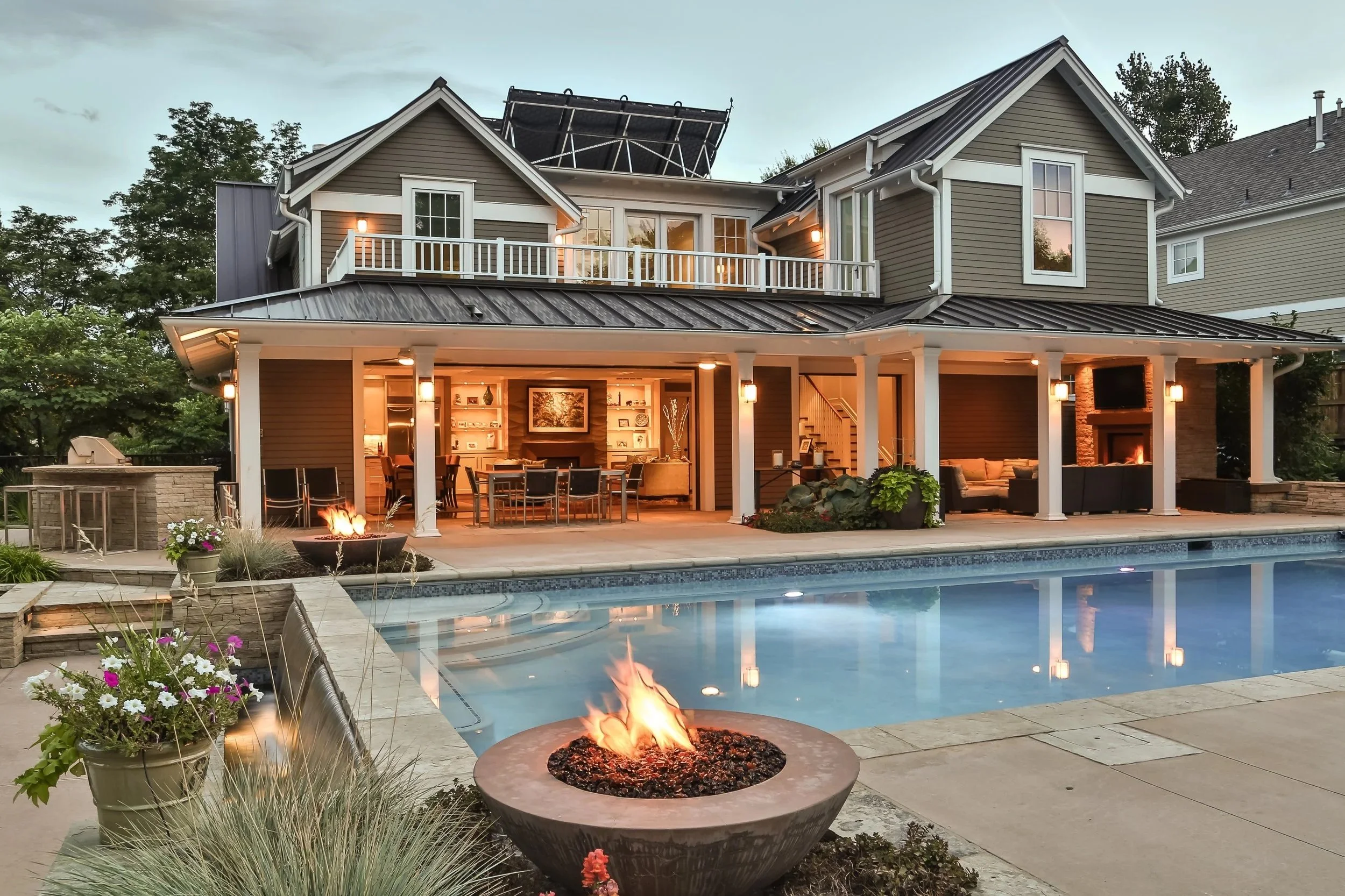 View of a modern two-story house with an outdoor living area, swimming pool, and fire pit, during evening hours. The house features a balcony, large windows, and a covered patio area illuminated with warm lighting. There's also a landscaped garden with flowers and trees around the pool area.