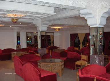Interior of a lounge with red upholstered chairs and sofas, a small wooden table with a candle, and decorative ceiling and floor columns, illuminated by warm lighting.