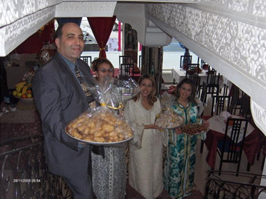 A group of three women and a man, all smiling and holding large trays of food, inside a restaurant or banquet hall with decorated windows and tables.