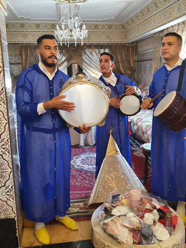 Three men dressed in blue traditional attire, playing drums indoors in a decorated room with a chandelier and patterned wallpaper.