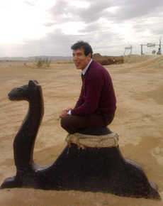 A man sitting cross-legged on top of a black camel-shaped sculpture in a desert landscape, with cloudy skies in the background.