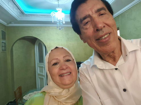 A man and woman smiling for a selfie indoors, with a chandelier and green walls in the background.