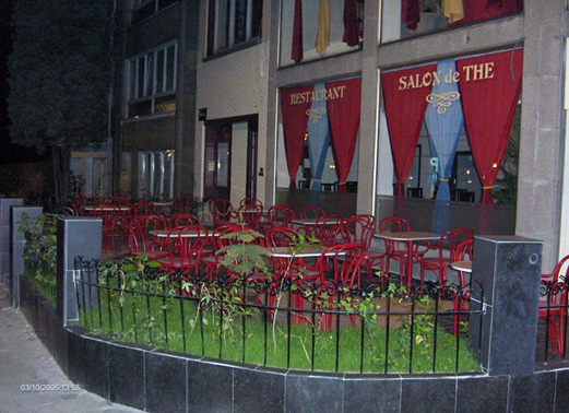 Outdoor seating area with red chairs and tables outside a restaurant with red curtains, located in an urban setting.