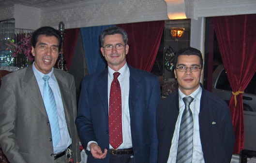 Three men in suits standing in front of curtains inside a room, smiling at the camera.