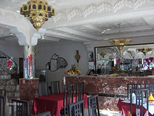 Interior of a restaurant with tables, chairs, a marble bar, ornate chandeliers, and decorative ceiling moldings.