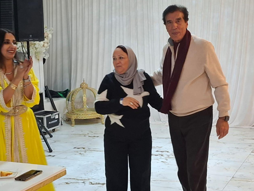 Three people at a celebration, two women and a man, standing in a decorated indoor space with white curtains. The woman on the left wears a yellow traditional dress, the woman in the middle wears a black outfit with a hijab, and the man on the right 