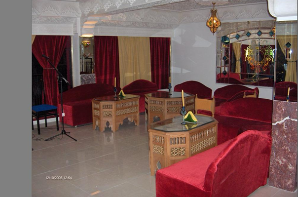 Interior of a lounge or waiting area with red velvet sofas, wooden tables, a microphone stand, and decorative curtains. Mirrors and ornate lighting fixtures are also visible.