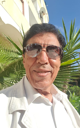 A man wearing sunglasses taking a selfie outdoors with a green plant and a building in the background.