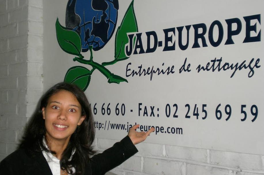 Young woman pointing to a sign for an cleaning company named JAD-EUROPE, with a globe and leaf logo, on a white brick wall.