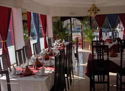 Interior of a restaurant with tables set for dining, white and pink curtains, and large windows with sunlight streaming in, decorated with potted plants and a chandelier.