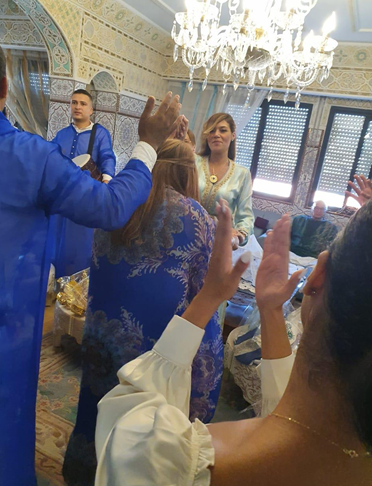 People gathered in an ornately decorated room, some raising their hands, with a woman in a light blue outfit standing in the center near a chandelier.