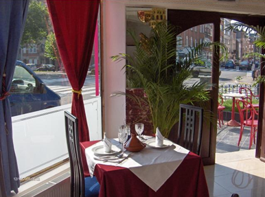 A dining table set for two in a restaurant with a red and white color theme, featuring a white tablecloth with a red overlay, glassware, napkins, and a brown vase. The restaurant has large windows, red curtains, and potted plants.