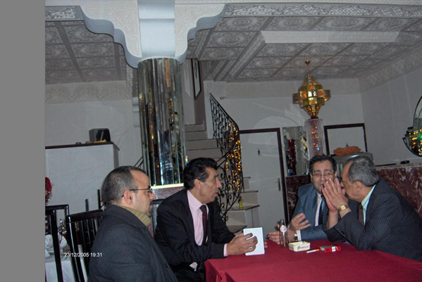 Four men sitting around a table having a conversation in a decorated room, with a staircase in the background.