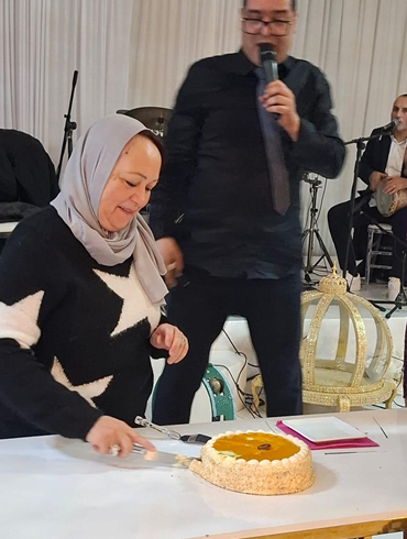 A woman wearing a hijab and a sweater with star pattern is cutting a cake on a table. A man standing behind her is holding a microphone, and in the background, there are musicians with instruments.