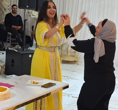 Two women dancing together at a festive event, one in a yellow dress and the other wearing a headscarf, with a DJ and equipment in the background.