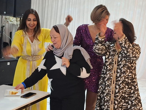 Four women smiling and dancing together indoors, one wearing a yellow dress, another in a hijab and black sweater, the third in a purple sequined dress, and the fourth in a leopard print outfit.
