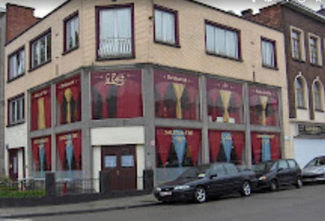 A corner of a multi-story building with large windows and red curtains, with cars parked in front on the street.