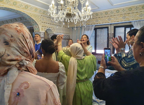 A group of women and men celebrating a special occasion in an elaborately decorated room with a chandelier and ornate walls, some taking photos while one woman appears to be singing or speaking.