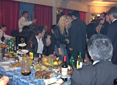 People gathered around a table with food and drinks at a social event, some standing and chatting.