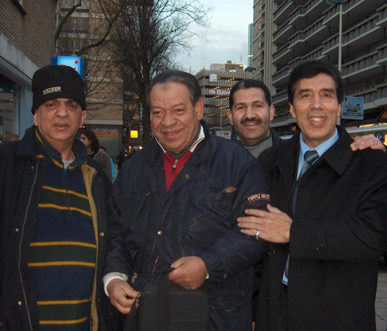 Four men standing together outdoors in an urban setting, smiling for the photo, with buildings and trees in the background.