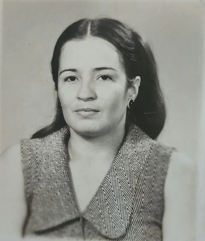 Black and white portrait of a woman with dark hair, wearing a sleeveless herringbone-patterned vest and small hoop earrings.