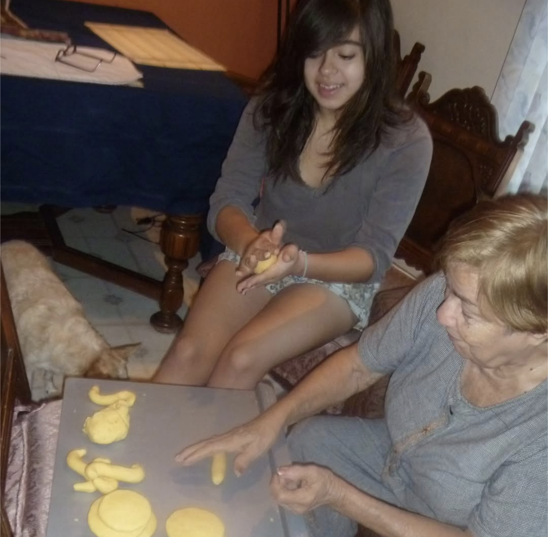 Two women preparing dough on a tray in a kitchen, with a dog on the floor near them waiting for crumbs.