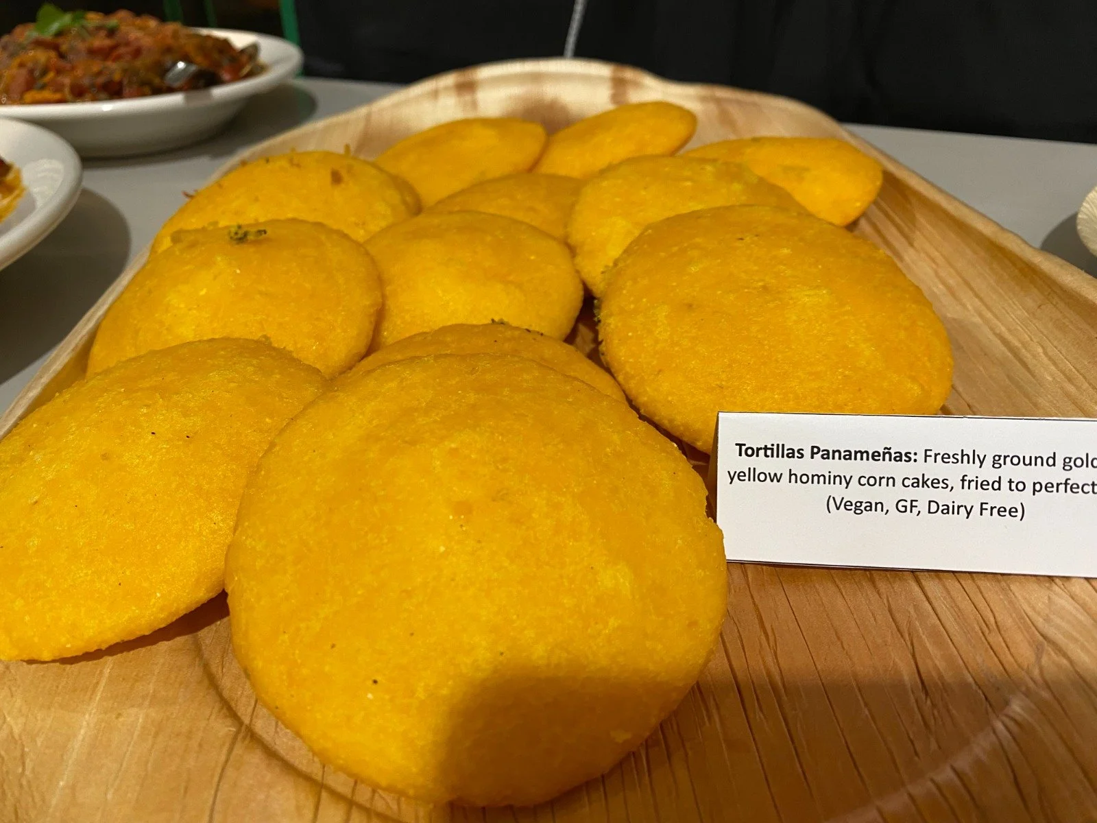 Several freshly ground yellow hominy corn cakes called Tortillas Panameñas on a wooden tray, fried to perfect golden color with a note indicating they are vegan, gluten-free, and dairy-free.