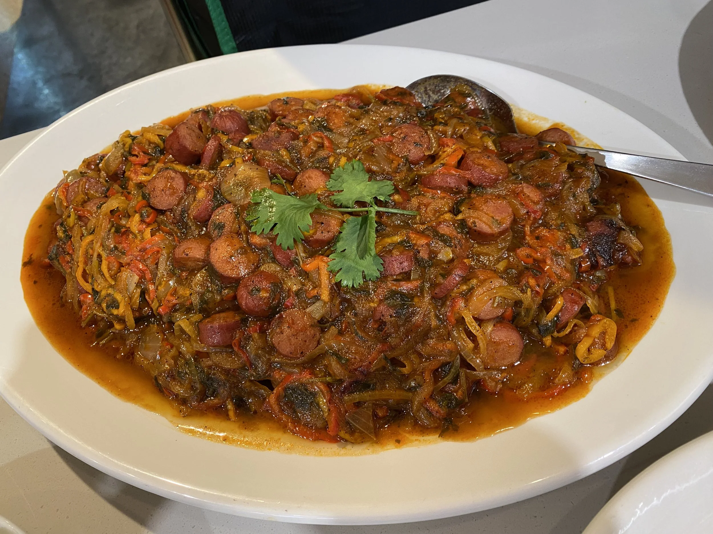 A white bowl filled with a spicy stew with sausage slices, vegetables, and garnished with cilantro, placed on a white surface.