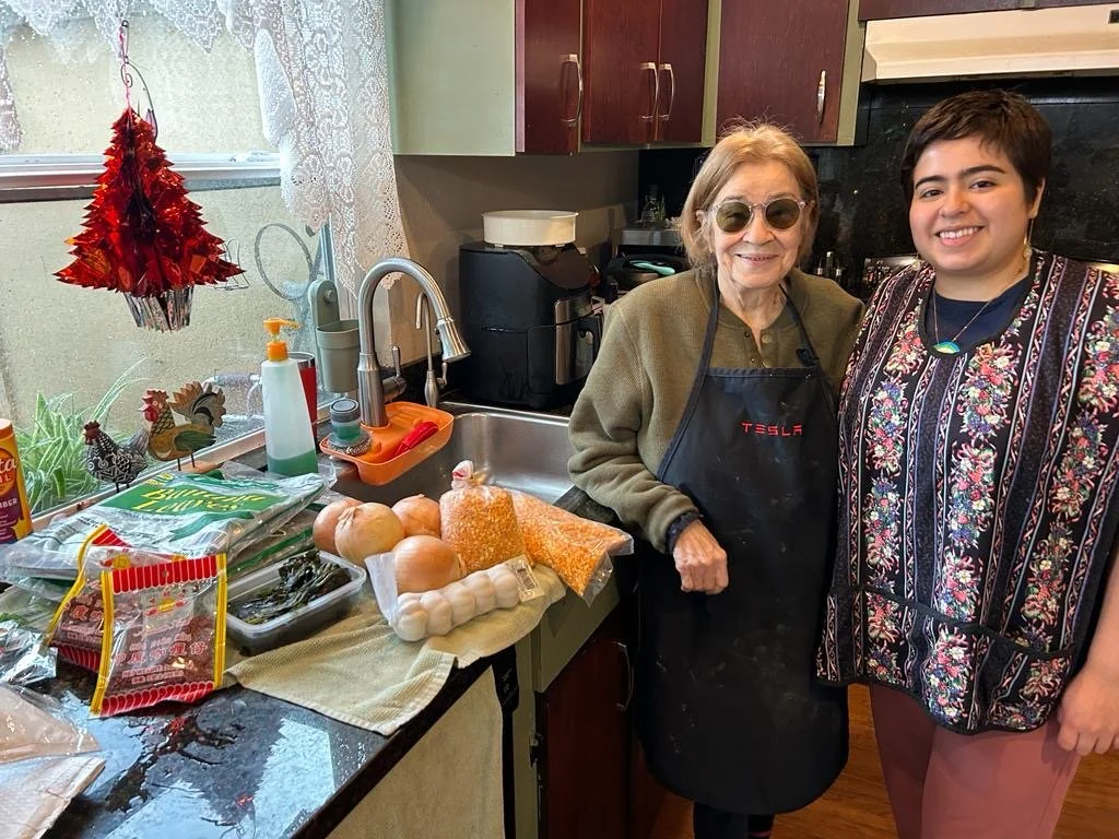 An elderly woman and a young woman both smiling in a kitchen. The elderly woman wears glasses and an apron that says 'TESLA', the young woman wears a floral top. On the kitchen counter are onions, garlic, a bag of rice, a bag of corn, and other cooking ingredients, with a window in the background.