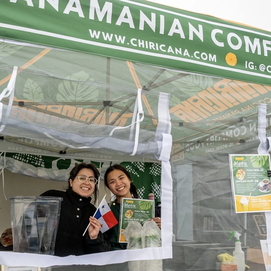 Two women smiling at a food stall under a green canopy with the sign 'Chiri Cana' and website 'www.chiricana.com'. One woman is holding a small Chilean flag. The stall offers dishes like Yuca Frita and Lentajas Plate.