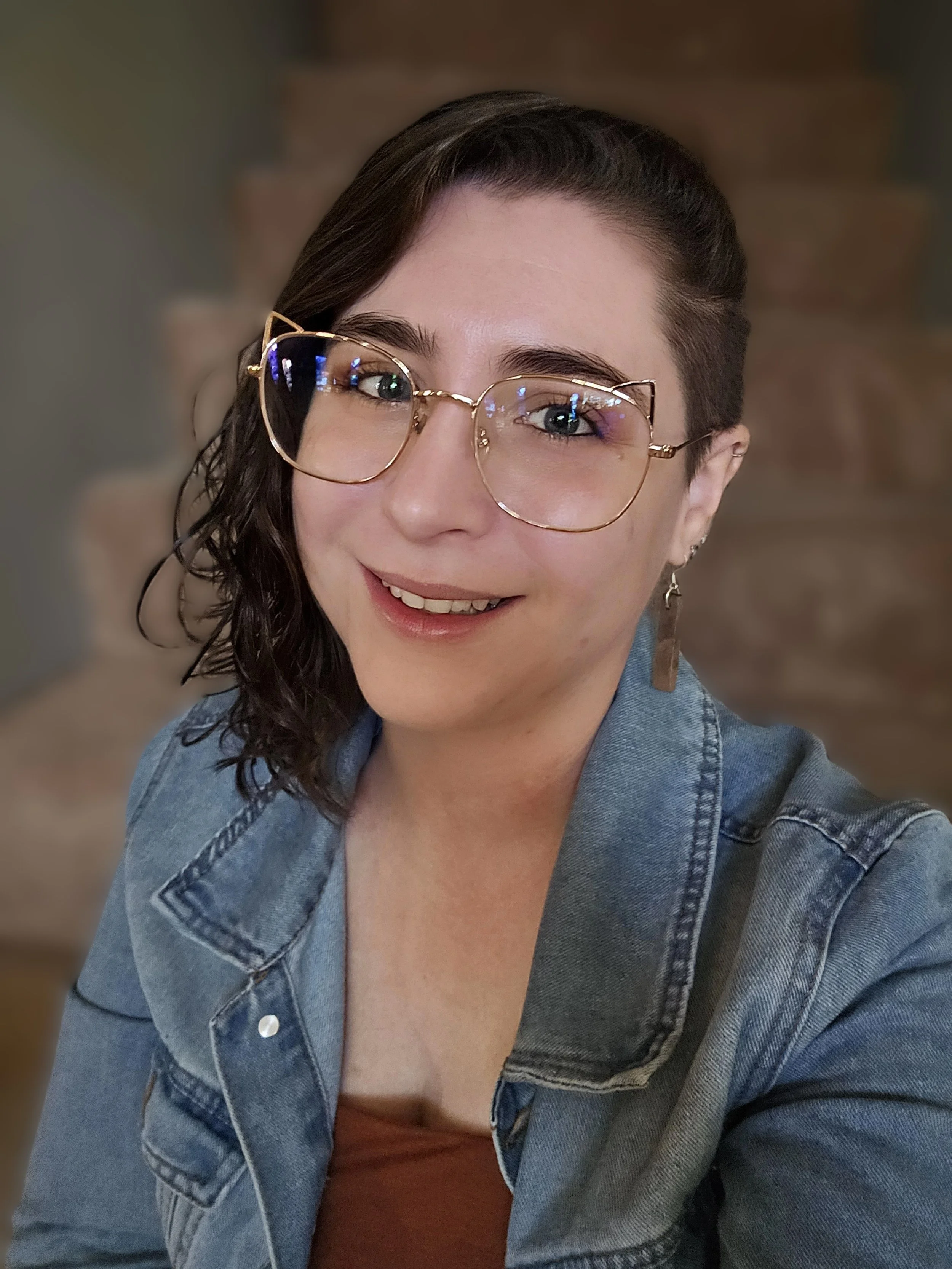 A woman with short dark hair and glasses smiling for a selfie, wearing a denim jacket and a brown top in front of a blurred background.