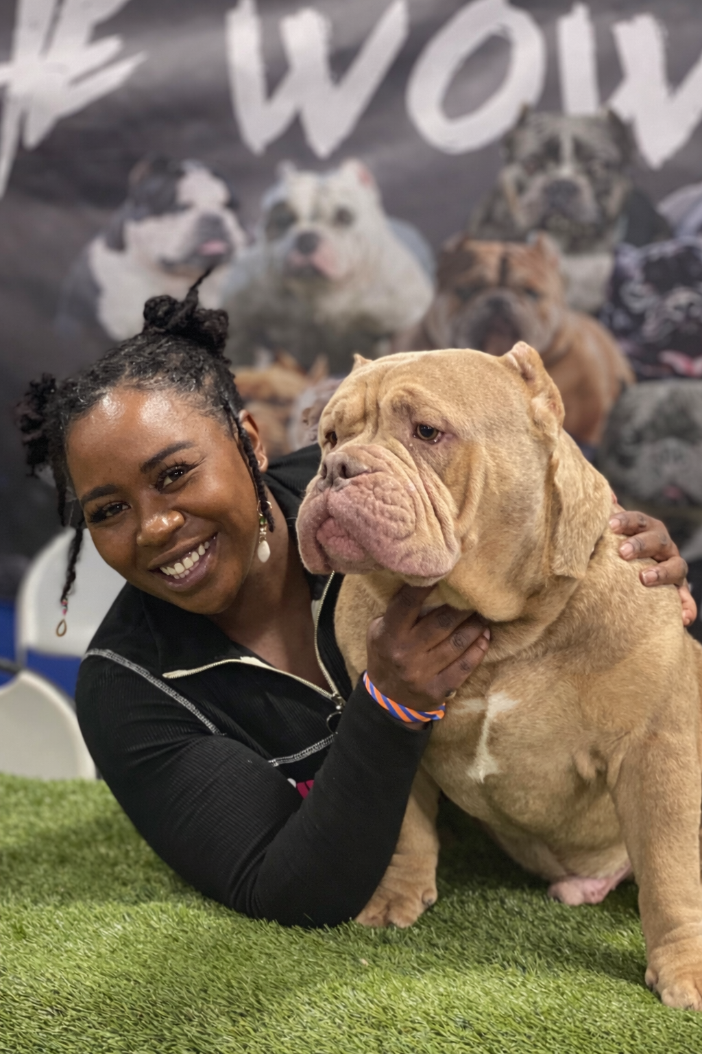A woman with dark skin, smiling and showing teeth, holding a large, light brown bulldog with a wrinkled face. They are on a green surface with a blurry background featuring various dog images.