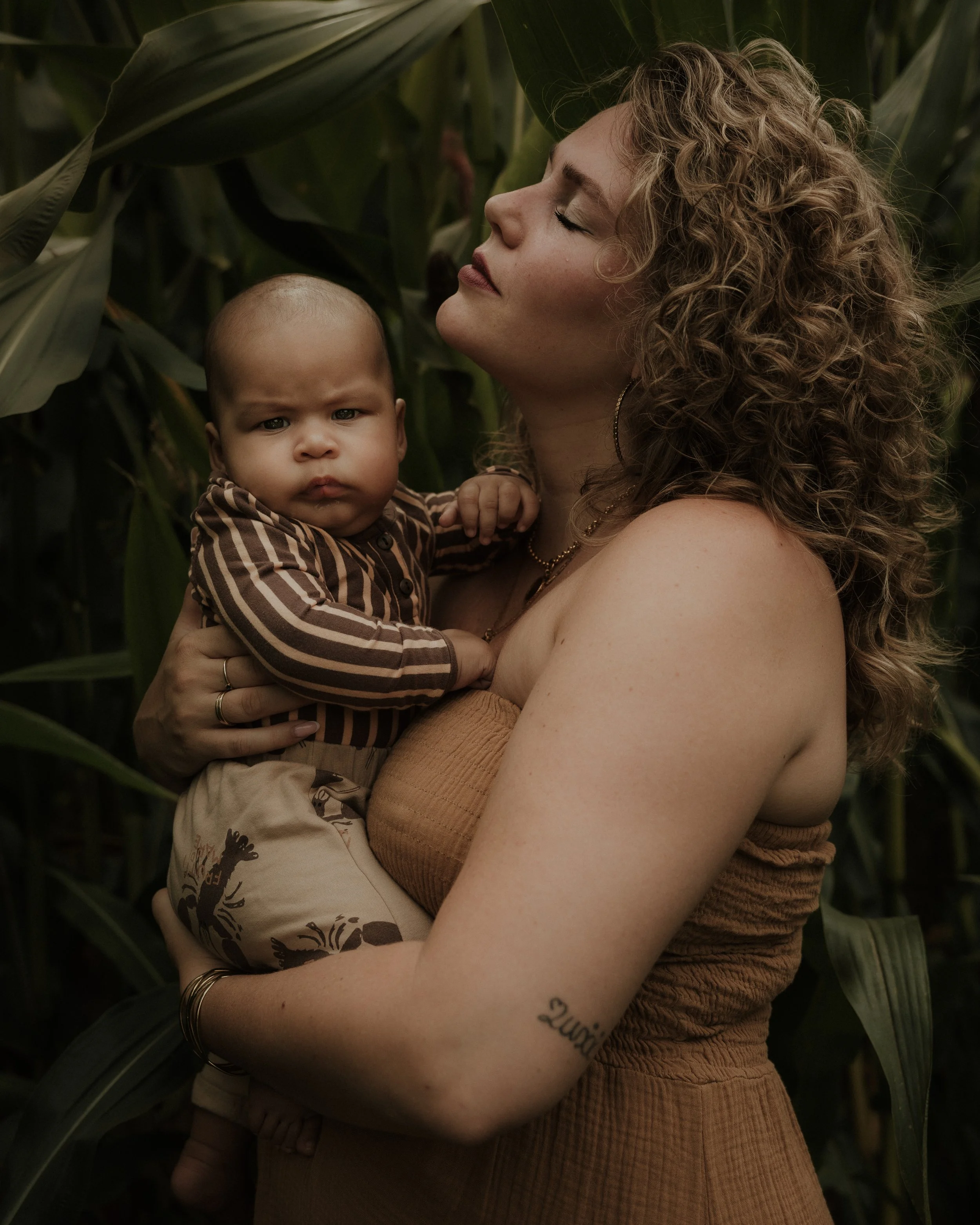 A woman with curly hair, wearing a strapless beige top and jewelry, holds a young child with a serious expression, wearing a striped brown and beige outfit, in a field of tall green plants.