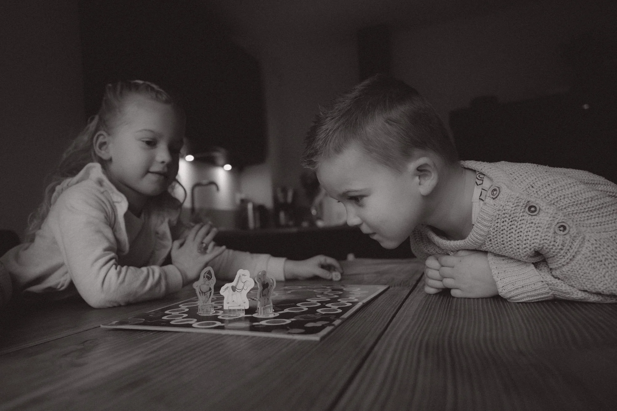 Two children, a girl and a boy, play a board game on a wooden table in a cozy room, with game pieces in the shape of horses.