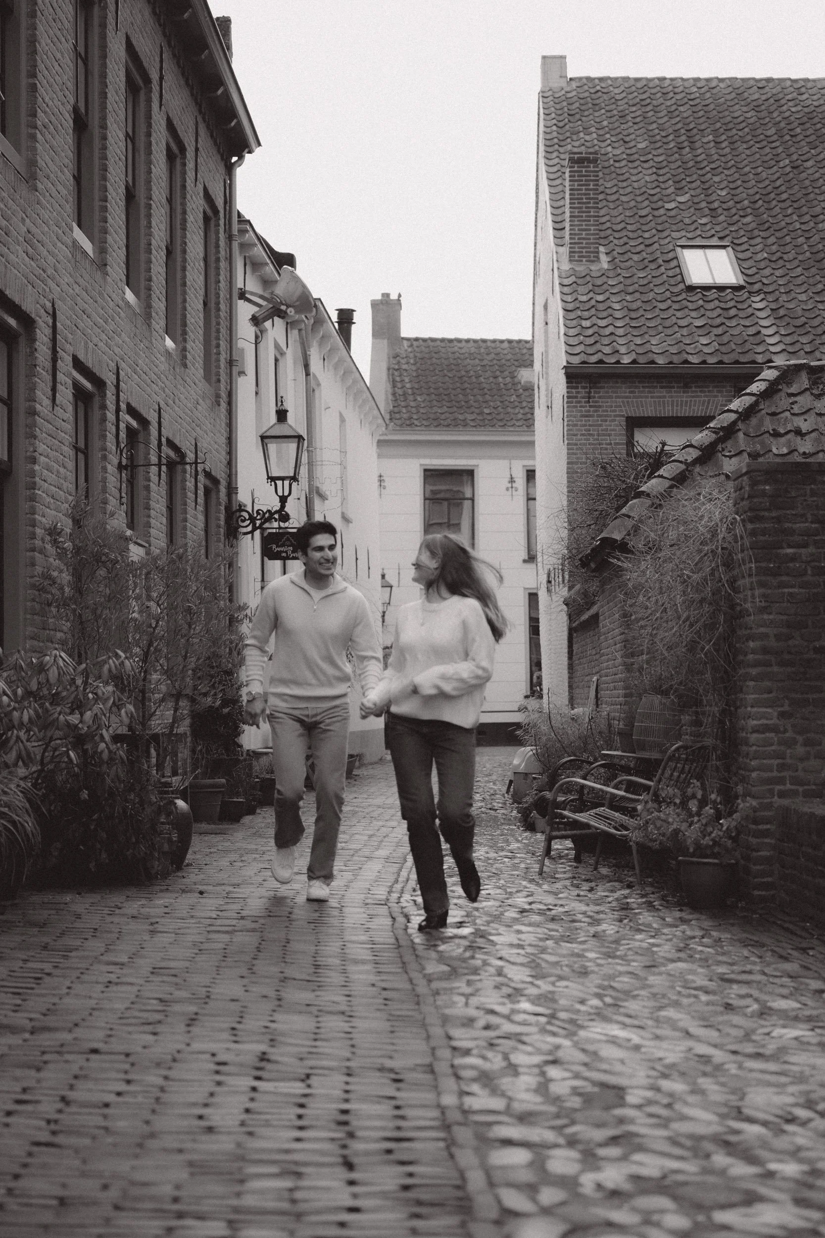A couple holding hands and running down a cobblestone alleyway in a European-style neighborhood, smiling and looking at each other, with old brick buildings and potted plants on the sides.